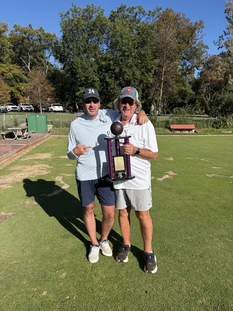 2026 club champion Mark Newton, holding the trophy, and Danny Newton stand arm in arm on the sunny green at Washington Park Lawn Bowling Club, both in caps and sunglasses, one pointing at the tall purple trophy topped with a bowl that the other holds, with big shade trees, a red bench and a green shed behind