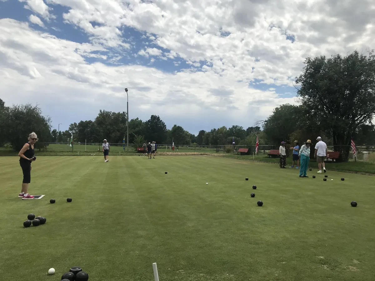 Bowlers spread across the wide Washington Park green on a bright day with billowing clouds, sets of black bowls scattered near the jack and flags along the far fence